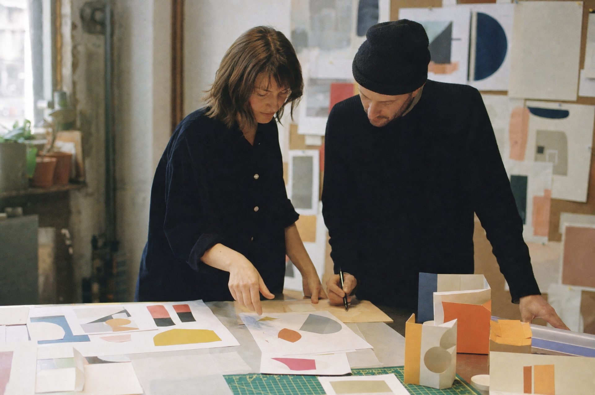 Two designers at a studio worktable covered with colour explorations and paper packaging mockups, one pointing at a sheet while the other takes notes